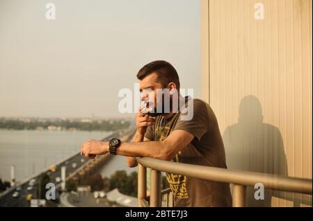 Getöntes Porträt von Hipster Kerl rauchen Zigarette. Ein Mann raucht auf dem Balkon. Ein Mann mit Bart steht auf einem Balkon hoch über einem Quadrat und raucht CI Stockfoto