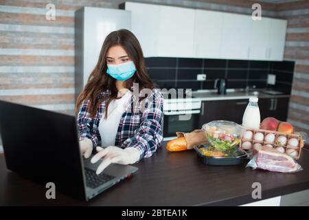 Konzept für die Lieferung von Lebensmitteln. Eine junge Frau in einer medizinischen Maske und einem Gummi-Einwegmaske Handschuhe bestellt Nahrung mit einem Laptop zu Hause. Auf dem Tisch sind Milch, Stockfoto