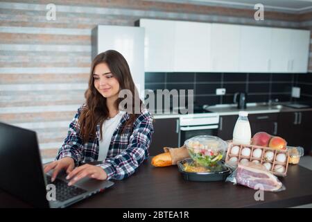 Konzept für die Lieferung von Lebensmitteln. Eine junge Frau bestellt zu Hause mit einem Laptop Essen. Auf dem Tisch stehen Milch, Salate in Kisten, Fleisch, Lebensmittel, Obst, Eier, Brot Stockfoto