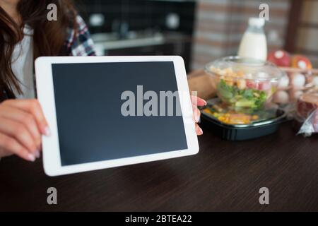 Konzept für die Lieferung von Lebensmitteln. Eine junge Frau bestellt zu Hause mit einem Laptop Essen. Auf dem Tisch stehen Milch, Salate in Kisten, Fleisch, Lebensmittel, Obst, Eier, Brot Stockfoto