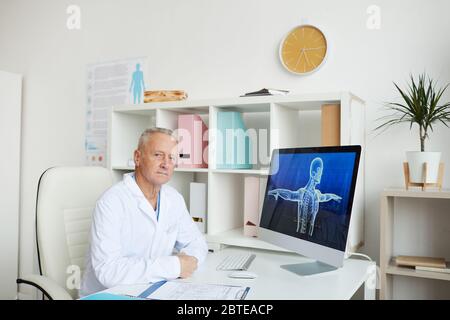 Porträt des modernen Senior Arzt sitzt am Schreibtisch im Büro und Blick auf die Kamera, mit digitalen Körper Scan-Bild auf dem Computer-Bildschirm, Kopierer Raum Stockfoto