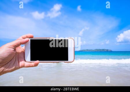 Tourist mit Telefon am Strand mit dem Meer, Hand halten weiße Handy Smartphone, Reise-Arbeitskonzept, verschwommener Hintergrund, Nahaufnahme. Stockfoto