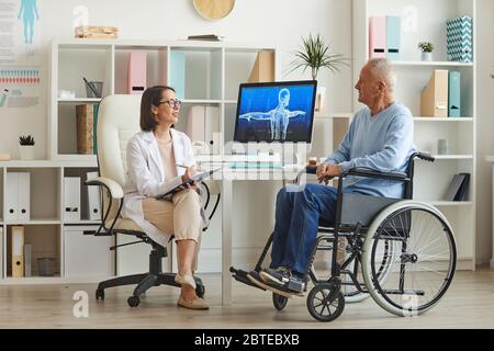 Ganzkörperporträt von behinderten älteren Mann im Rollstuhl im Gespräch mit Ärztin während der Konsultation in der Klinik Stockfoto