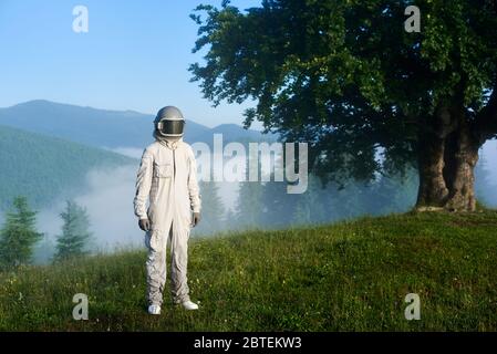 Porträt eines Raumfahrers mit weißem Raumanzug und Helm auf einer grünen Bergwiese auf einem Hügel, großem alten Baum und fantastischem Nebelfichtenwald in einem Tal auf dem Hintergrund. Reisekonzept Stockfoto