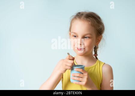 Nettes kleines Mädchen mit lustigen Zöpfen hält Plastikbecher mit Joghurt und einem Teelöffel Vorbereitung zu essen. Nahaufnahme Studio-Porträt isoliert auf hellblau Stockfoto