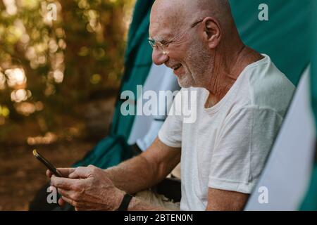 Lächelnder älterer Mann, der mit seinem Mobiltelefon im Zelt sitzt. Pensionierter Mann, der in der Natur campiert und SMS auf seinem Handy liest. Stockfoto