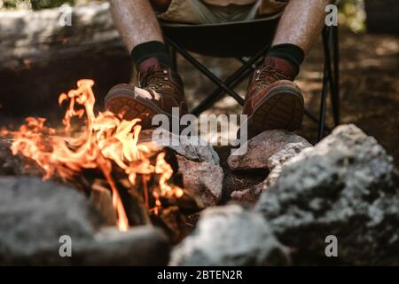 Mann in Stiefeln wärmt sich am Feuer auf dem Campingplatz die Füße. Kurzer Schuss eines älteren Mannes, der am Lagerfeuer sitzt. Stockfoto