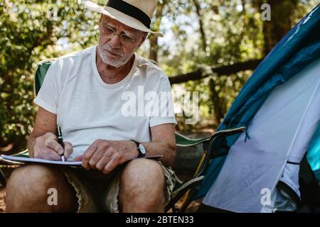 Älterer Mann, der vor einem Zelt sitzt und in einem Buch schreibt. Älterer kaukasischer Mann, der ein Buch auf dem Campingplatz schreibt. Stockfoto