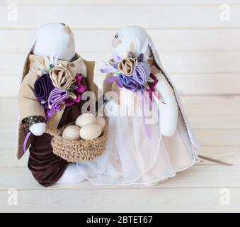 Zwei handgemachte Kaninchen halten einen Korb mit Eiern. Weißer Holzhintergrund. Beige und lila. Stockfoto