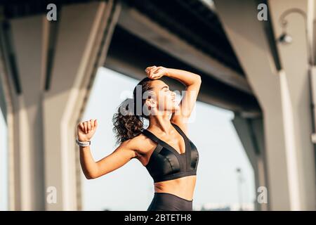 Seitenansicht der Frau in Fitness trägt Strecken ihre Arme. Lächelnde Sportlerin, die sich vor dem Training im Freien aufwärmt. Stockfoto