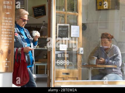 Ältere Frau, die Kaffee schlürft, steht am Eingang zu einem netten Café in Ericeira, Portugal, eine junge Frau sitzt an einem Tisch in einem großen Fenster Stockfoto