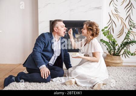 Frisch vermählte. Frau füttert ihren Mann mit einem Kuchen, während sie auf dem Teppich vor dem Kamin drinnen sitzt Stockfoto