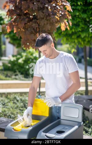 Dunkelhaariger Mann in Schutzhandschuhen, der Plastikbeutel in den Abfallbehälter wirft Stockfoto