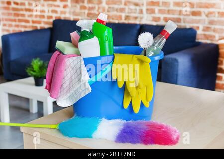 Eimer mit Schwämmen, Waschmittelflaschen und Wischstock vor dem Sofa im Wohnzimmer. Reinigungsgeräte für die Hausarbeit. Idee für Reinigungsservice. Stockfoto