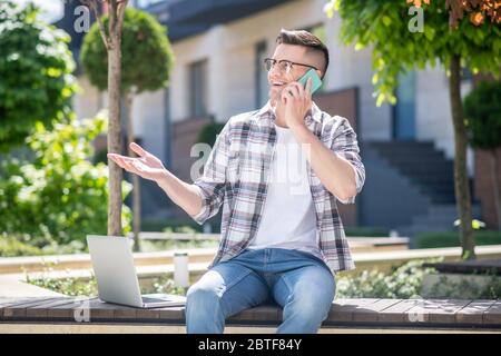 Dunkelhaariger Mann mit Brille, sitzt mit seinem Laptop im Hof und spricht mit seinem Handy Stockfoto