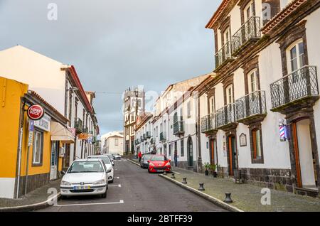Lagoa, Azoren, Portugal - 13. Jan 2020: Stadtstraße im Fischerdorf Lagoa. Geparkte Autos auf der Asphaltstraße, traditionelle Gebäude und Kirche im Hintergrund. Horizontales Foto. Stockfoto