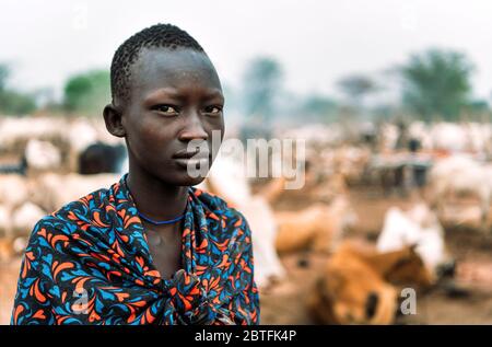 MUNDARI TRIBE, SÜDSUDAN - 11. MÄRZ 2020: Teenager in traditionellem farbenfrohem Outfit, der die Kamera gegen die verschwommene ländliche Umgebung im Süden betrachtet Stockfoto