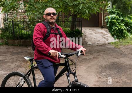 Ein älterer Mann mit grauem Bart und Glatze in Brille und einem roten Pullover mit Kapuze auf einer Schlange sitzt auf seinem Mountainbike. Gesunder Sport und Leben Stockfoto