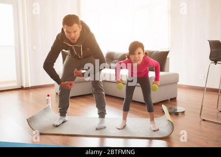 Vater und Tochter trainieren zu Hause. Training in der Wohnung. Sport zu Hause. Sie machen Gesichter und haben Spaß auf einer Yogamatte mit Hanteln Stockfoto