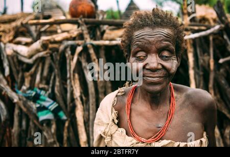 TOPOSA STAMM, SÜDSUDAN - 12. MÄRZ 2020: Alte blinde Frau aus dem Toposa Stamm auf verschwommenem Hintergrund der Stockhütte im Dorf im Südsudan Stockfoto