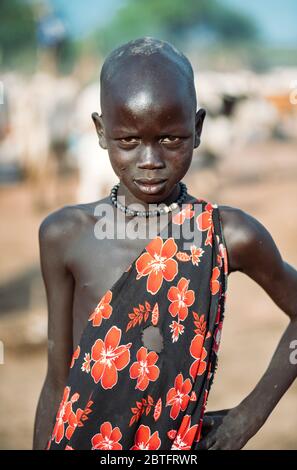 MUNDARI TRIBE, SÜDSUDAN - 11. MÄRZ 2020: Kind in traditionellem Gewand mit floralem Ornament, das die Hand auf der Taille hält und während des Lebens die Kamera betrachtet Stockfoto