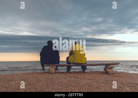 Zwei Männer sitzen auf einer Bank in der Nähe eines Strandes und schauen mit epischen Wolken auf das Meer. Stockfoto