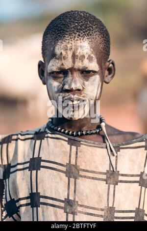 MUNDARI STAMM, SÜDSUDAN - 11. MÄRZ 2020: Teenager in traditionellen Gewand und mit Gesicht mit Schlamm bemalt während der Leben in Mundari Tribe Dorf in Stockfoto