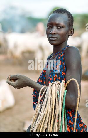 MUNDARI TRIBE, SÜDSUDAN - 11. MÄRZ 2020: Teenager in traditioneller Kleidung, die Seile trägt und die Kamera betrachtet, während sie in der Nähe von Rindern auf der Weide arbeiten Stockfoto