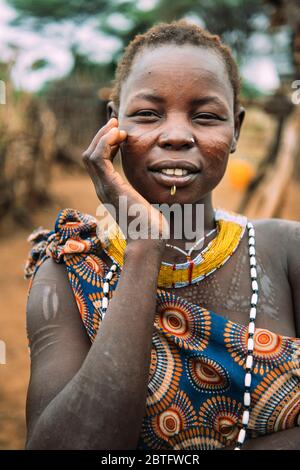 TOPOSA STAMM, SÜDSUDAN - 12. MÄRZ 2020: Junge Frau mit traditionellen Narben des Toposa Tribe berühren Gesicht und Blick auf Kamera auf verschwommen Stockfoto