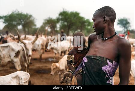 MUNDARI STAMM, SÜDSUDAN - 11. MÄRZ 2020: Frau in traditioneller Kleidung und Accessoires, die neugieriges Baby an den Händen trägt, während sie sich dagegen stellt Stockfoto