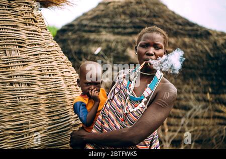 TOPOSA STAMM, SÜDSUDAN - 12. MÄRZ 2020: Frau aus Toposa Stamm Pfeife rauchen und Baby tragen, während in der Nähe von traditionellen Hütten im Dorf in Stockfoto