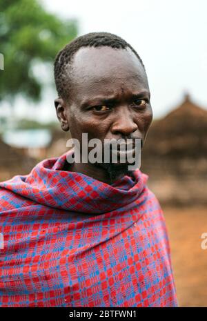 TOPOSA STAMM, SÜDSUDAN - 12. MÄRZ 2020: Erwachsener Mann in kariertem Tuch schaut Kamera an, während sie auf verschwommenem Hintergrund des Toposa Tribe steht Stockfoto