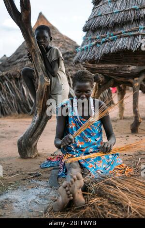 TOPOSA STAMM, SÜDSUDAN - 12. MÄRZ 2020: Frau sitzt auf schmutzigem Boden in der Nähe von Hütte und Kind und Weberei Produkt aus Stroh im Dorf Toposa Tribe Stockfoto