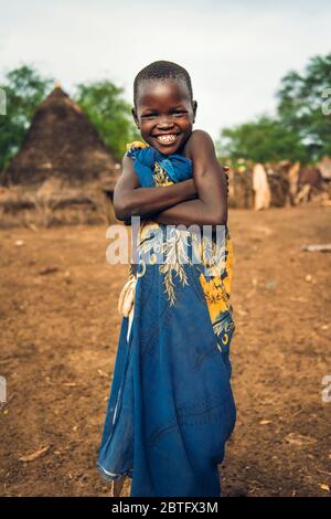 TOPOSA STAMM, SÜDSUDAN - 12. MÄRZ 2020: Glückliches Kind in buntes Tuch eingehüllt lächelnd für Kamera und Arme kreuzen während des Lebens in Toposa Tribe Stockfoto