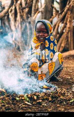 TOPOSA STAMM, SÜDSUDAN - 12. MÄRZ 2020: Kind in ornamentalen Tuch lächelnd und schaut Kamera, während sie auf Hacken in der Nähe von kleiner Flamme sitzt Stockfoto