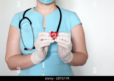 Frau Ärztin in medizinischer Maske, die rotes Strickherz in den Händen hält und während einer COVID-19-Coronavirus-Pandemie Leben rettet. Kardiologe, Blutspender Stockfoto