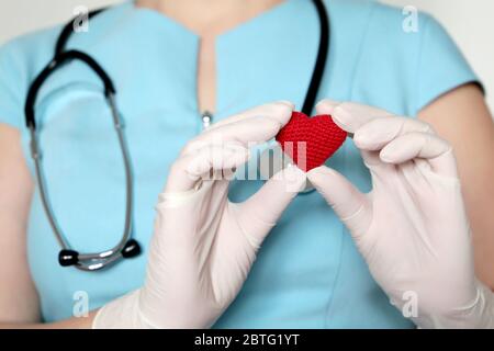 Frau Ärztin in medizinischer Maske, die rotes Strickherz in den Händen hält und während einer COVID-19-Coronavirus-Pandemie Leben rettet. Kardiologe, Blutspender Stockfoto