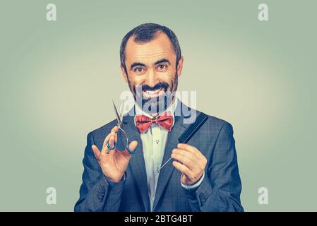 Lächelnder bärtiger Friseur mit eleganter Jacke mit roter Fliege, posiert mit Schere und Kamm. Stilvolle Erwachsene Mann Studio Porträt, isoliert bei gre Stockfoto