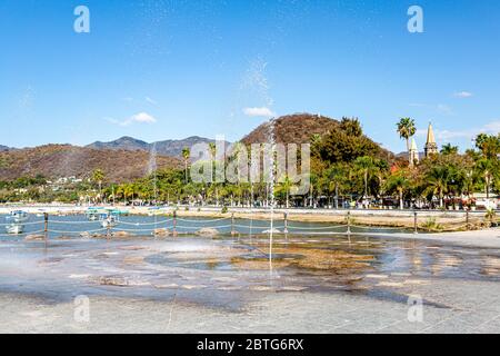 Brunnen, der den Boden mit Wasserstrahlen auf der Promenade des Sees Chapala mit der Stadt im Hintergrund, sonnigen Tag mit einem klaren blauen Himmel Stockfoto