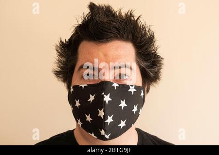 Ein Mann in den späten Dreißigern mit einer wiederverwendbaren Gesichtsmaske, der während der Coronavirus-Pandemie Covid 19 mit verrücktem, absperrbarem Haar direkt auf den Betrachter blickt Stockfoto