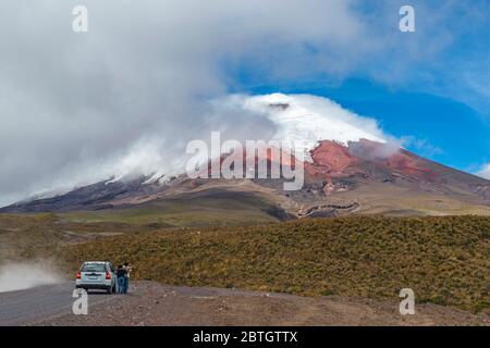 Touristen halten an einer unbefestigten Straße mit ihrem Fahrzeug, um den Blick über den schneebedeckten Gipfel des aktiven Vulkans Cotopaxi, Quito, Ecuador, zu genießen. Stockfoto