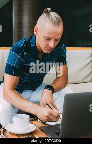 Ein junger Mann schreibt auf ein Notizbuch, nachdem er die Post auf einem Laptop überprüft hat, arbeitet ferngesteuert in einem Café über das Internet. Tasse Kaffee neben einem offenen Laptop, ser Stockfoto