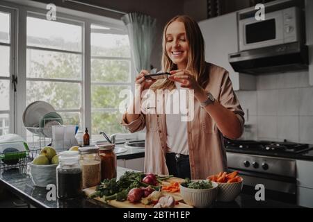 Junge Frau, die während der Isolation und Quarantäne in der modernen Küche zuhause mit dem Smartphone von ihrem Essen fotografiert Stockfoto