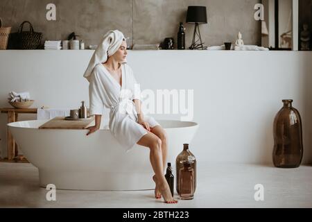 Attraktive junge Frau im Bademantel sitzt auf dem Rand der Badewanne.