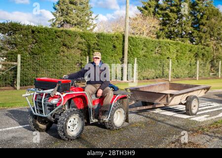 Annat, Canterbury, Neuseeland, Mai 26 2020: Ein pensionierter Bauer schlemmt einen Anhänger hinter seinem Quad, um Heckenscheren auf einer Landstraße zu sammeln Stockfoto