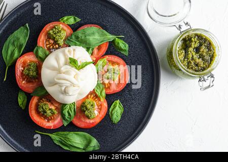 Burrata Büffelkäse mit frischen Tomaten und Basilikumblättern Pesto-Sauce auf schwarzem Teller weißen Hintergrund Flatlay serviert. Stockfoto