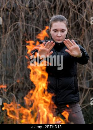 Ein junges Mädchen in einer schwarzen Jacke, das seine Hände ausstreckt, wärmt seine Handflächen am Feuer. Leuchtend orange Lagerfeuer Flammen. Verschwommener Hintergrund trockener Äste. Stockfoto