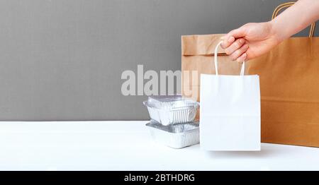 Weiße Handtasche aus Bastelpapier in weiblicher Hand. Lebensmittelfolienbehälter und Papierverpackungen auf grauem Tischhintergrund. Lieferservice für Speisen. Essen zum Mitnehmen. Kopieren Stockfoto