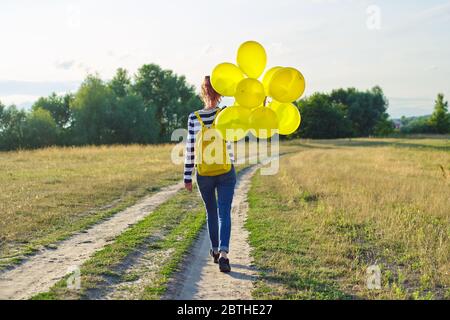 Teenager-Mädchen mit Rucksack und gelben Ballons auf Landstraße, Rückansicht Stockfoto