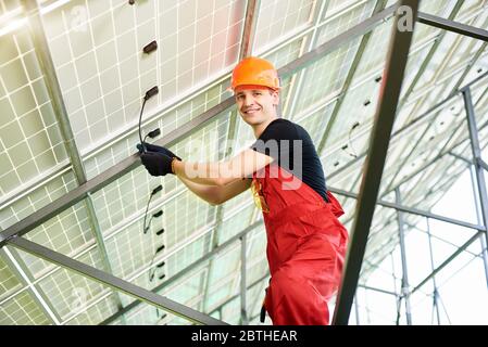 Männlicher Ingenieur, der hochwertige Solarmodule inspiziert. Arbeiter in orangefarbener Uniform und Helm mit Ausrüstung, lächelnd zur Kamera. Photovoltaik Stromversorgungssysteme. Konzept für grüne Energie. Stockfoto